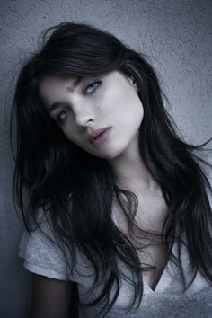 a woman with long black hair and blue eyes posing for a photo in front of a gray wall