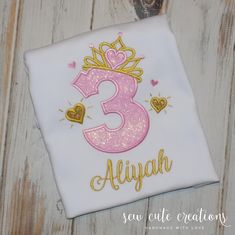 a pink and gold birthday shirt with the number three on it's chest, sitting on a wooden surface