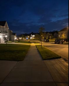 an empty street at night with cars parked on the side and houses in the background