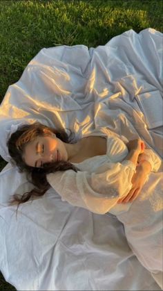 a young woman laying on top of a white blanket in the grass with her eyes closed