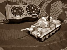 a toy tank sitting on top of a bed next to a remote control controller and an alarm clock