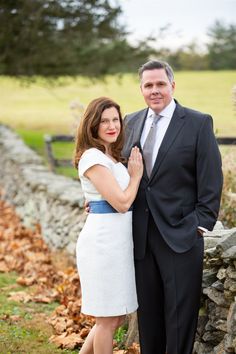 a man and woman standing next to each other in front of a stone wall with leaves on the ground