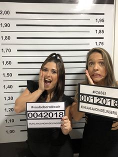 two women holding up signs in front of a mugshot wall with numbers on them