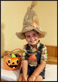 a young boy sitting on top of a bed wearing a scarecrow hat and holding a pumpkin