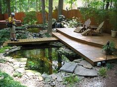 a wooden deck over a small pond surrounded by rocks and plants with chairs around it