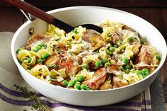 a white bowl filled with pasta and peas on top of a table next to a wooden spoon