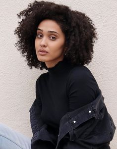 a woman with curly hair sitting on the ground wearing jeans and a black shirt is looking at the camera