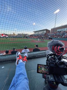 a person with their feet on a camera in front of a baseball field and net