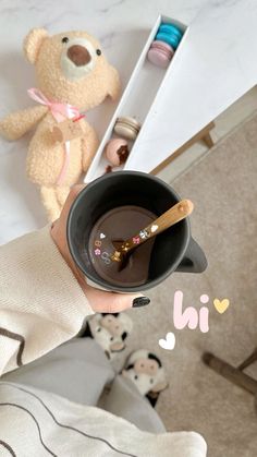 a teddy bear sitting on the floor next to a person holding a cup with chocolate in it