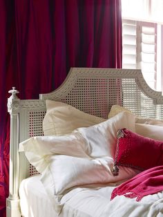 a white bed with red pillows and blankets on it's headboard next to a window