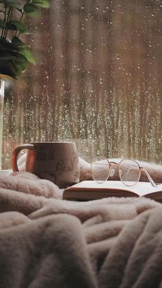 an open book, coffee cup and glasses sitting on a bed in front of a rainy window