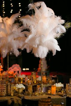 there is a tall centerpiece with white feathers on top of gold plates and glasses