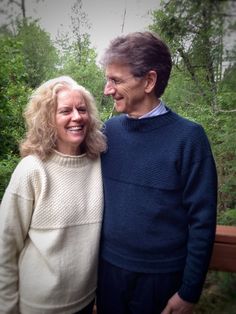 an older couple standing next to each other in front of some trees and bushes, smiling for the camera