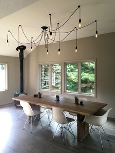 a dining room table with white chairs and lights hanging from the ceiling above it in front of two windows