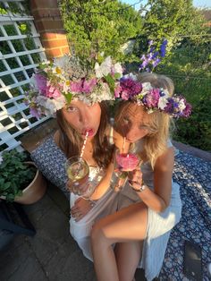 two women sitting on a bench with drinks in their hands and flower crowns over their heads