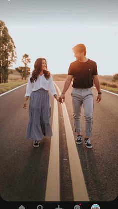 a man and woman holding hands walking down the middle of an empty road with trees in the background