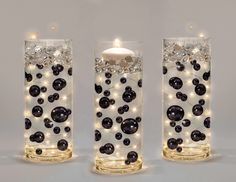 three glass vases with lights in them on a white surface, one is filled with black balls