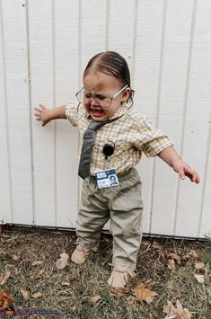 a toddler wearing glasses and a tie standing in front of a white wall with leaves on the ground