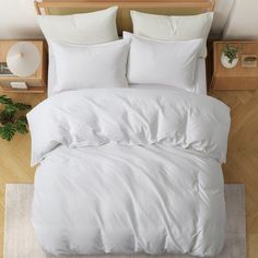 a bed with white sheets and pillows on top of a wooden floor next to a plant