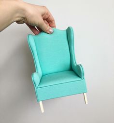 a hand holding a miniature blue chair on a white wall with nails sticking out of it