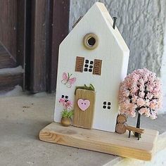 a small white house sitting on top of a wooden stand next to a tree and flowers