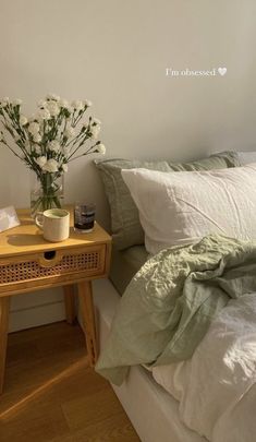 a small wooden table with flowers on it next to a white bed and green sheets