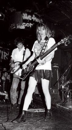 a woman playing guitar while standing next to another person on stage with other people behind her