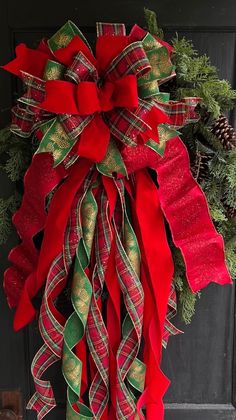 a red and green christmas wreath hanging on the front door with pine cones, evergreens and ribbons