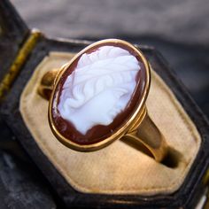 This storied ring showcases an oval hardstone cameo of a person wearing a headband, possibly laurel leaves. There are slight nicks on the hardstone, but nothing distracting. The ring is crafted in 10k yellow gold which we have left unpolished, and is currently a size 6.5. Luxury Vintage Cameo Signet Ring, Timeless Oval Intaglio Signet Ring, Timeless Oval Signet Ring With Intaglio, Timeless Oval Intaglio Ring, Timeless Oval Ring With Intaglio Detail, Luxury Cameo Ring As Gift, Classic Oval Carved Ring, Classic Carved Oval Ring, Timeless Oval Intaglio Jewelry