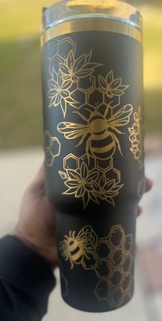 a person holding up a black and gold tumbler cup with bees on the side