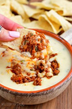 a hand holding a tortilla chip over a bowl filled with cheese and ground beef