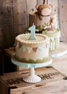 two tiered cakes decorated with teddy bears and one number on top, sitting on wooden crates