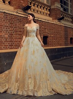 a woman in a gold and white wedding dress standing on the sidewalk next to a brick building