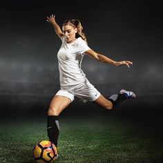 a woman kicking a soccer ball on top of a green field with lights in the background