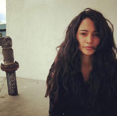 a woman with long hair standing next to a fire hydrant and looking at the camera