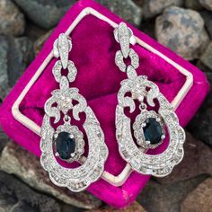 This gorgeous pair of earrings each feature a centered dangling oval mixed cut natural sapphire, four-prong set, in a halo of twelve (12) round brilliant cut diamonds. The dangle suspends from a fleur-de-lis adorned frame accented with twenty-nine (29) round brilliant cut diamonds and edged with twenty-two (22) baguette cut diamonds. The earrings measures approximately 35.42mm long and 15.60mm wide. The earrings are finished with friction backs for pierced ears. We have the matching necklace under SKU A95218. Oval Sapphire Earrings With 17 Jewels, Luxury Oval Sapphire Diamond Earrings, Luxury Oval Sapphire Earrings, Elegant Oval Diamond Earrings With 17 Jewels, Formal Sapphire Diamond Earrings With Diamond Accents, Exquisite Oval Diamond Earrings With Accents, Classic Oval Sapphire Diamond Earrings, Elegant Sapphire Earrings With Diamond Cut, Vintage Oval Diamond Earrings For Formal Occasions