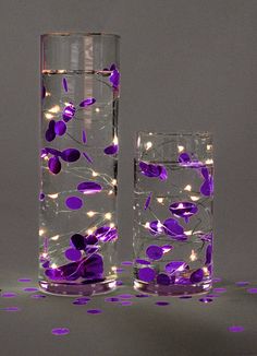 two tall vases with purple flowers and lights on the inside one is filled with water