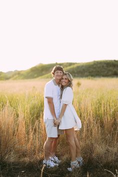 a man and woman standing in the middle of a field with tall grass behind them