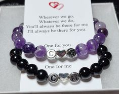 two bracelets with purple and black beads are on display in a white card box