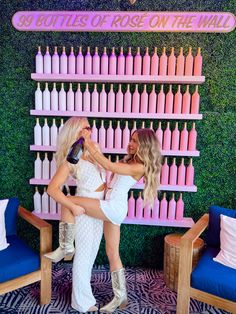 two women standing next to each other in front of a wall with bottles on it