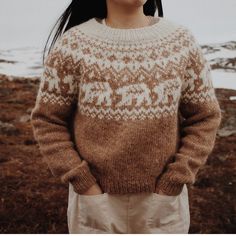 a woman wearing a brown and white sweater standing in front of snow covered mountains with her hands in her pockets