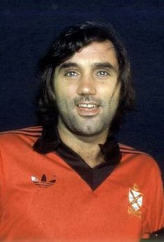 a man with long hair wearing an orange shirt and looking at the camera while standing in front of a dark background