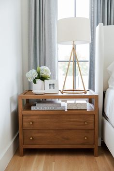 a nightstand with books and flowers on it in front of a window next to a bed