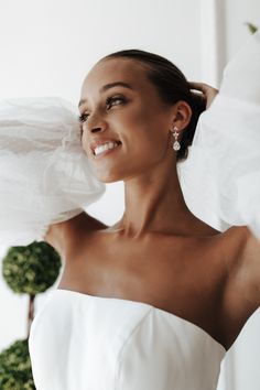 a woman in a white dress is smiling and holding her veil over her head with one hand