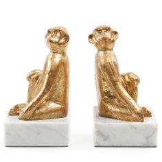 two golden monkey figurines sitting on white marble bases, one with its head in the other's hands