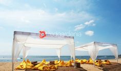 two white canopy beds sitting on top of a sandy beach next to the ocean under a blue sky