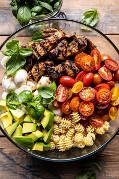 a glass bowl filled with pasta, tomatoes and chicken meats next to fresh basil leaves