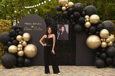 a woman standing in front of a black and gold birthday backdrop with balloons on it