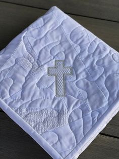 a white quilted blanket with a cross embroidered on the front and bottom, sitting on a wooden surface