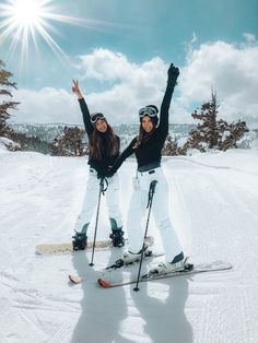 two women standing on skis in the snow with their arms up and hands raised